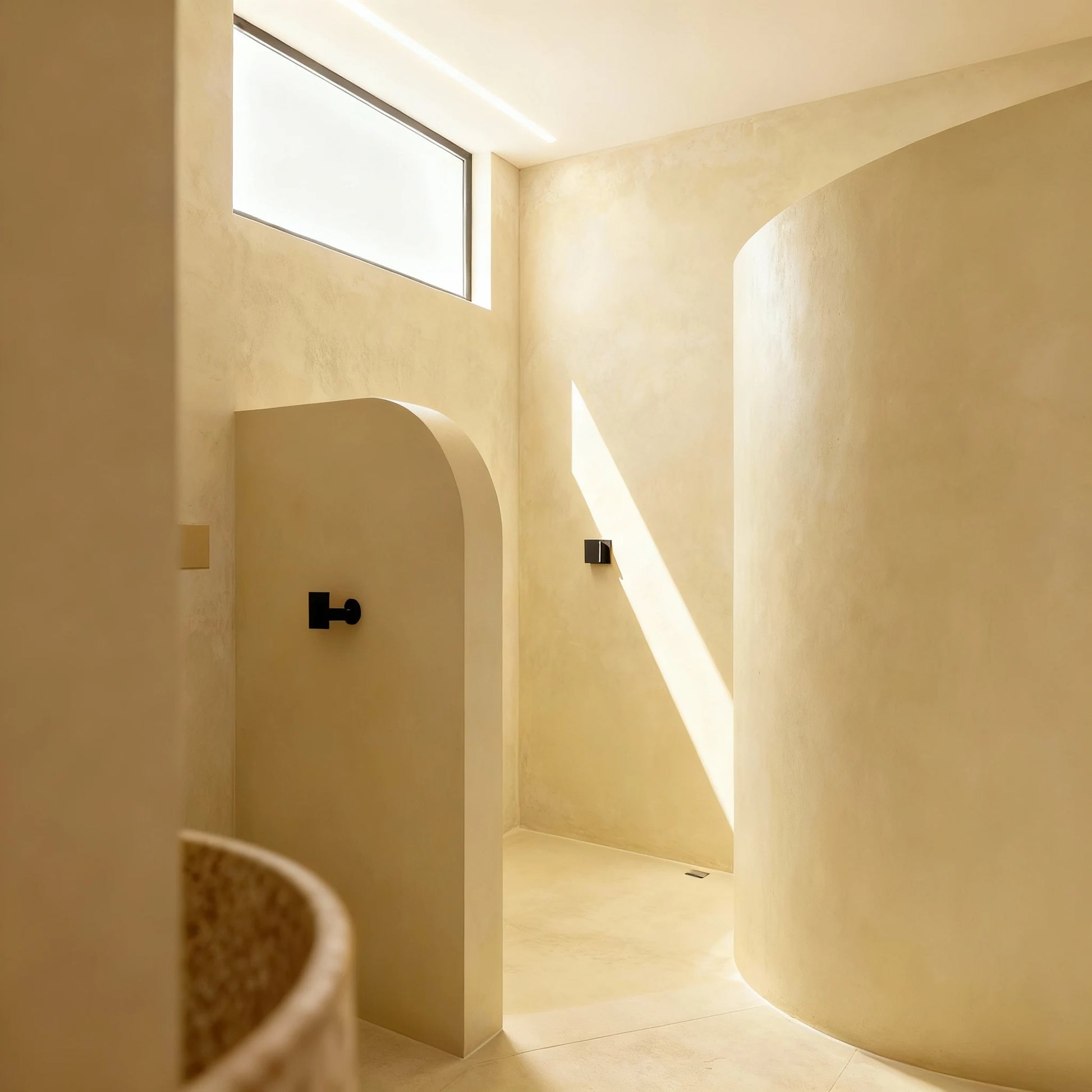 Ducha de obra en microcemento color India, paredes curvas y luz natural que entra por una ventana alta.