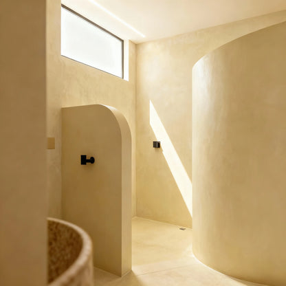 Ducha de obra en microcemento color India, paredes curvas y luz natural que entra por una ventana alta.