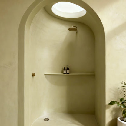 Ducha de obra con arco revestida con microcemento color Khaki, luz cenital suave y repisa integrada con frascos de baño.