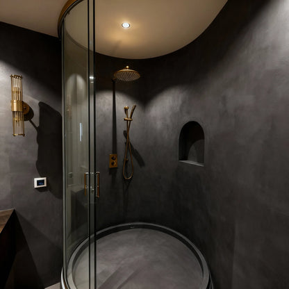 Original ducha de obra circular con microcemento color Shale Grey, grifería dorada y luz ambiental cálida.