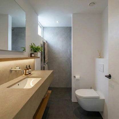 Baño estrecho con encimera de microcemento marrón color Mojave, lavabo integrado, inodoro suspendido y contraste de suelo oscuro con paredes claras.
