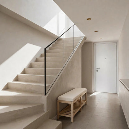 Recibidor de vivienda con escalera revestida en microcemento beige color Bone, barandilla de cristal, banco de madera y luz natural.