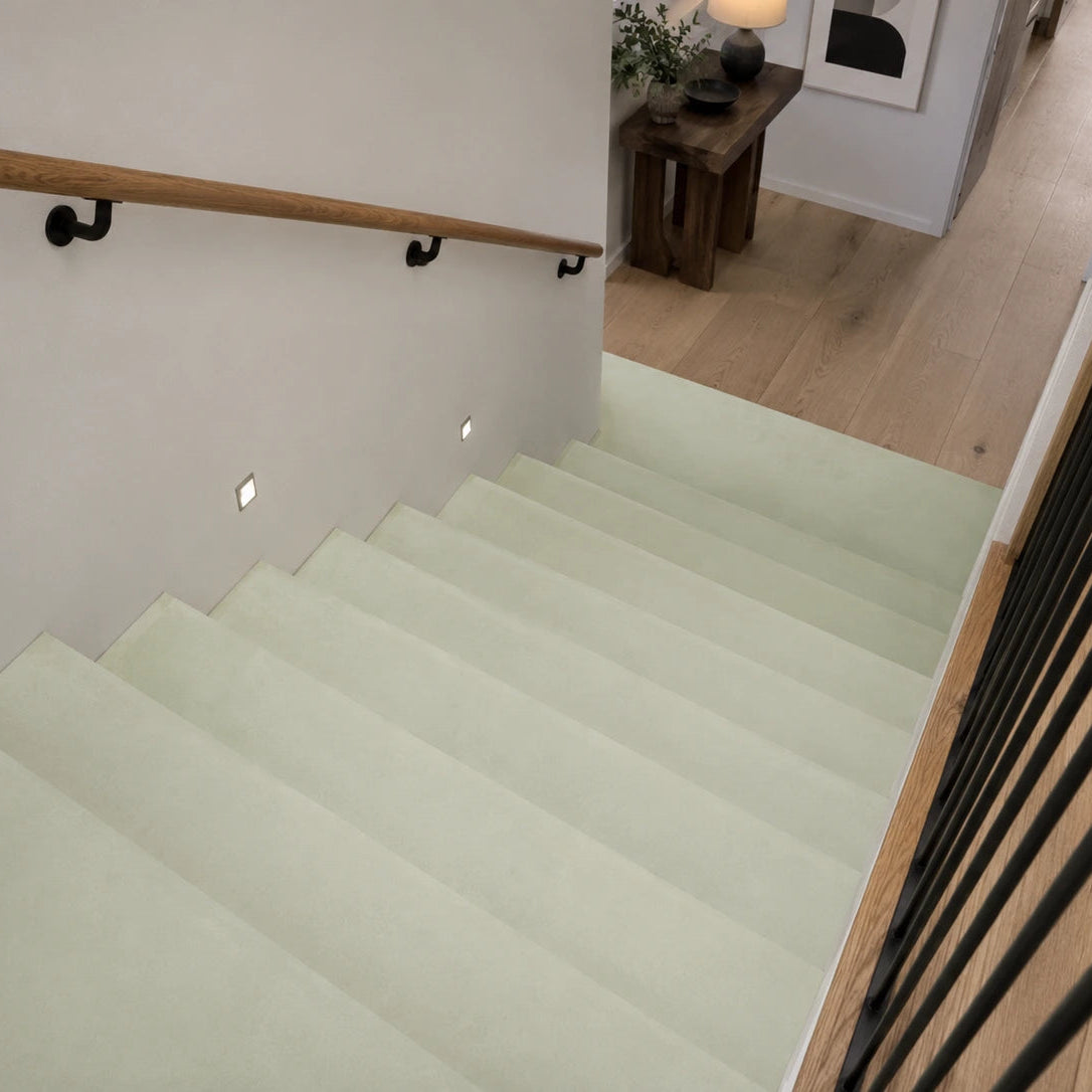 Tramo de escalera interior revestida en microcemento verde suave color Moss, con acabado continuo mate, pasamanos negro y transición a suelo de parquet.