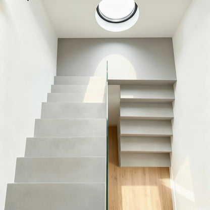 Escalera de microcemento color Liquen con líneas rectas y barandilla de cristal, iluminada por una ventana circular superior.