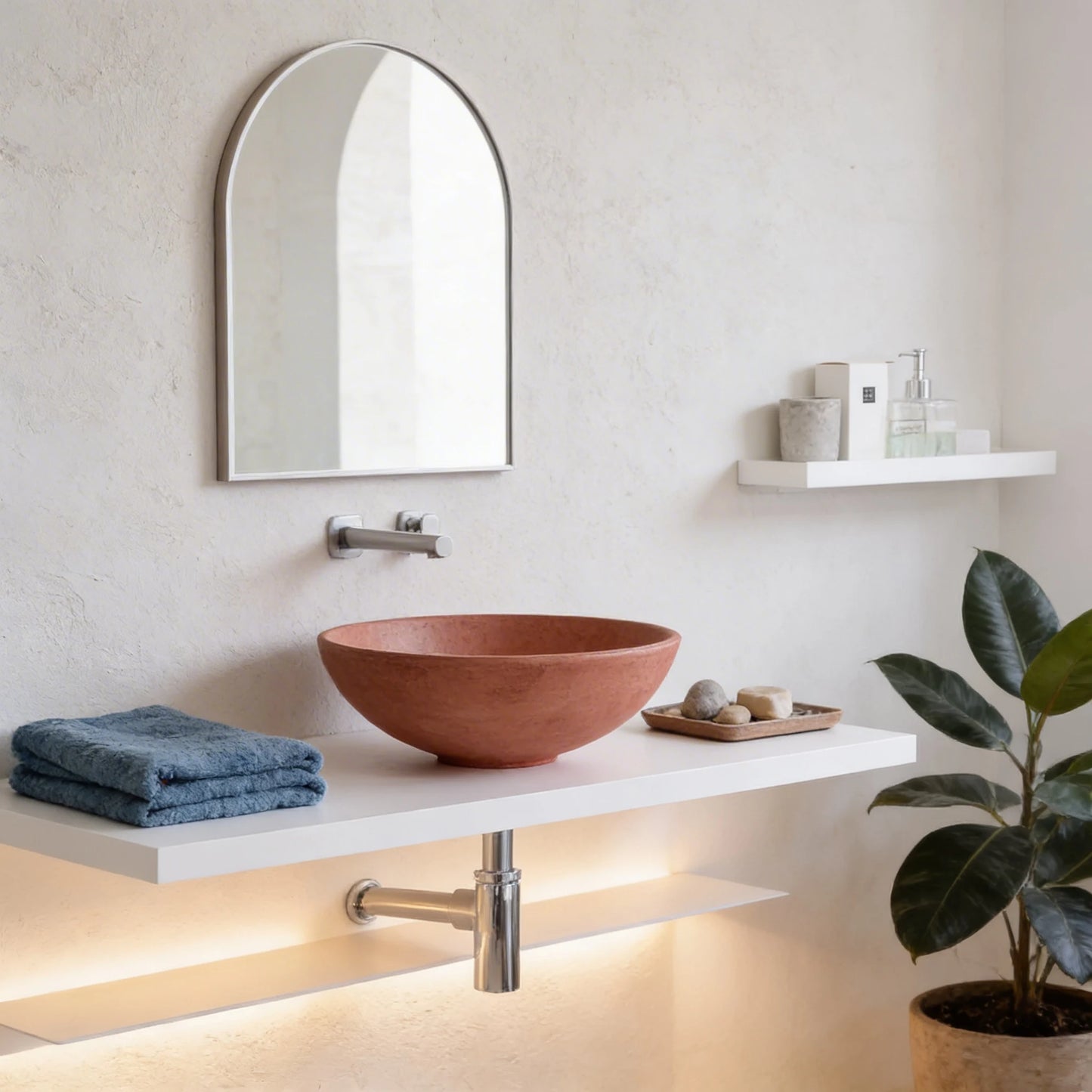 Lavabo redondo de microcemento en color Coral sobre encimera blanca flotante, con grifería mural cromada, espejo arqueado y decoración minimalista en baño moderno.