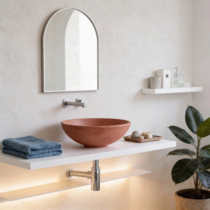 Lavabo redondo de microcemento en color Coral sobre encimera blanca flotante, con grifería mural cromada, espejo arqueado y decoración minimalista en baño moderno.