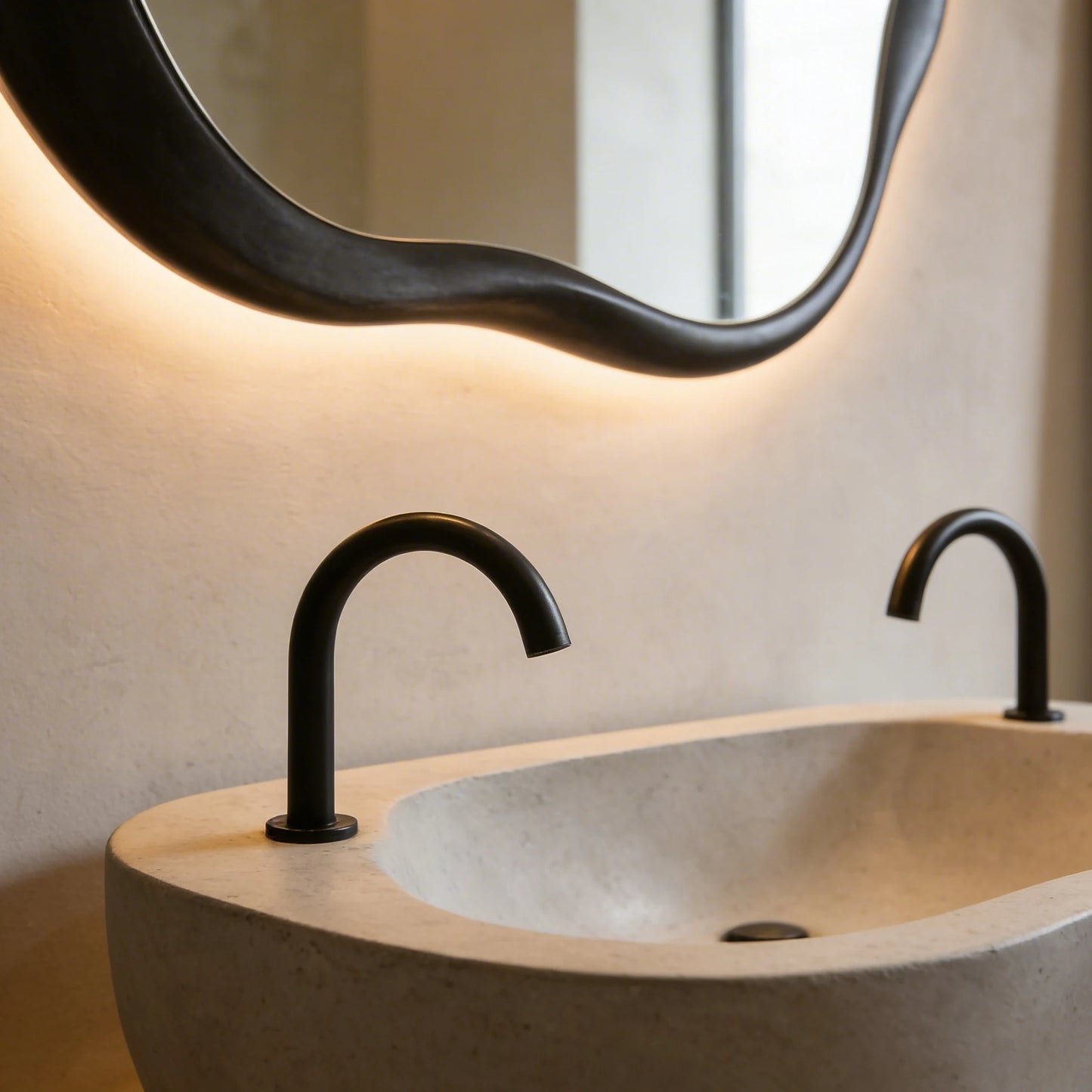 Lavabo de microcemento de formas orgánicas en color Bone, con acabado liso y textura mineral suave, grifería negra mate y espejo irregular retroiluminado sobre pared neutra.