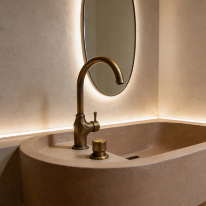 Lavabo de microcemento color Café  con forma ovalada y bordes redondeados, grifería en acabado latón envejecido y espejo redondo con iluminación indirecta.