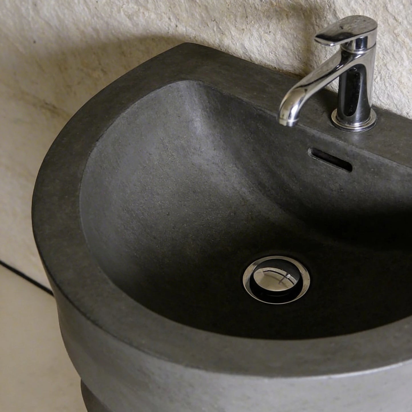 Lavabo redondo de microcemento en color Shale Grey con acabado mineral, grifería cromada y desagüe metálico, visto en primer plano sobre pared texturizada clara.