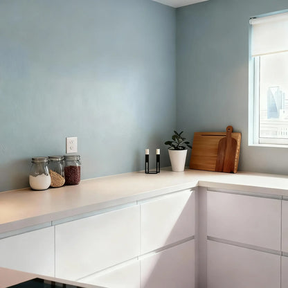 Pared de cocina en microcemento color Breeze con encimera blanca, tarros de ingredientes y tablas de madera junto a la ventana.