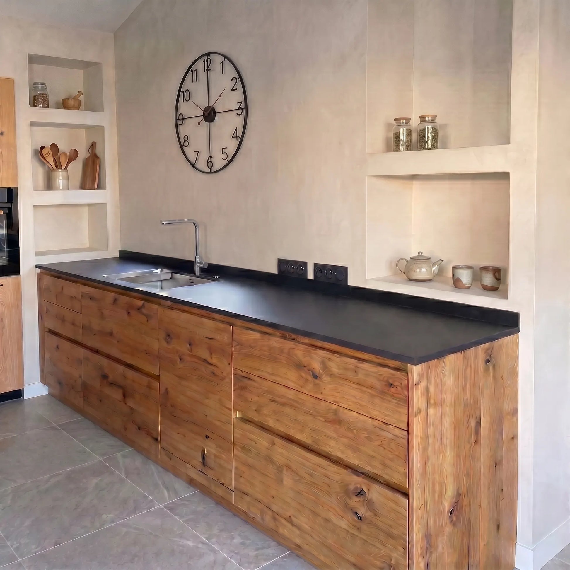 Cocina con paredes de microcemento Smartcret color Porcelain, muebles de madera natural, encimera negra con fregadero integrado y nichos decorativos.