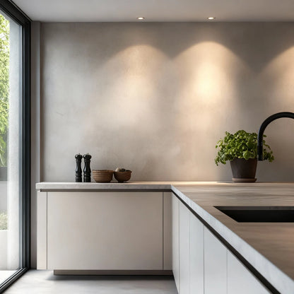 Pared de cocina en microcemento color Papyrus con encimera clara, iluminación superior y maceta verde junto a la zona del fregadero.