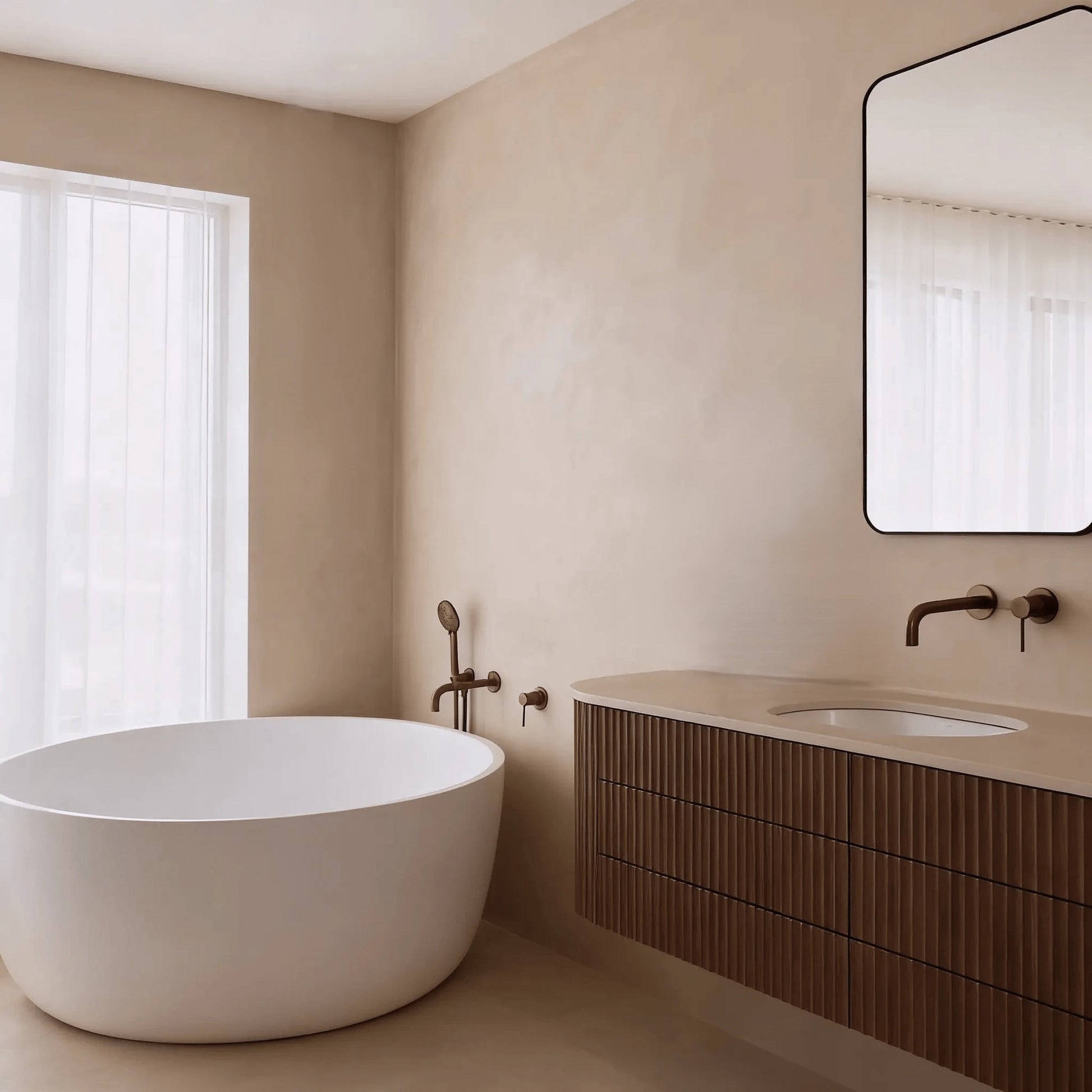 Pared de baño en microcemento color Bone con bañera exenta blanca, mueble estriado oscuro y luz suave filtrada.