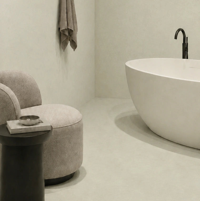 Microcemento gris color Liquen en suelo de baño minimalista con bañera blanca, sillón tapizado, iluminación indirecta y tonos suaves.
