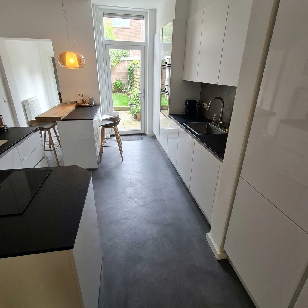 Cocina estrecha y alargada con pavimento de microcemento gris oscuro color Shale Grey, mobiliario blanco, encimera negra y puerta acristalada al exterior.