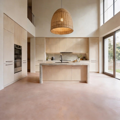 Cocina abierta con isla y suelo de microcemento rosa empolvado color Blush de Smartcret, gran ventanal y lámpara de mimbre suspendida.