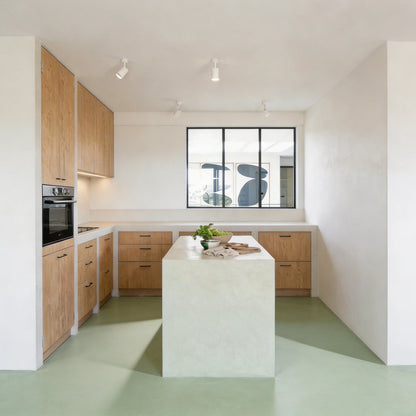 Cocina de estilo mediterráneo con suelo continuo de mcirocemento verde color Moss de Smartcret, isla central revestida en blanco y mobiliario de madera clara.